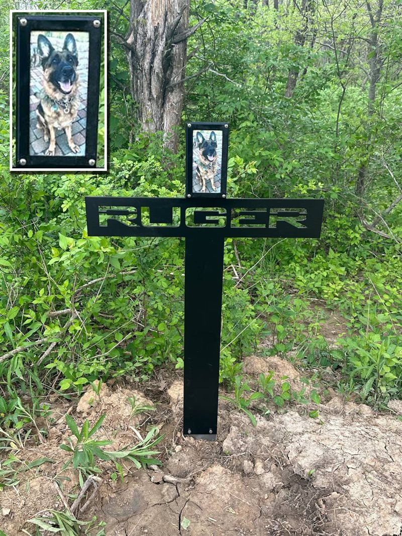 Black steel memorial cross cut with the name Ruger, holding a photo of a German Shepherd, set among green trees, with a close-up of his photo in the corner